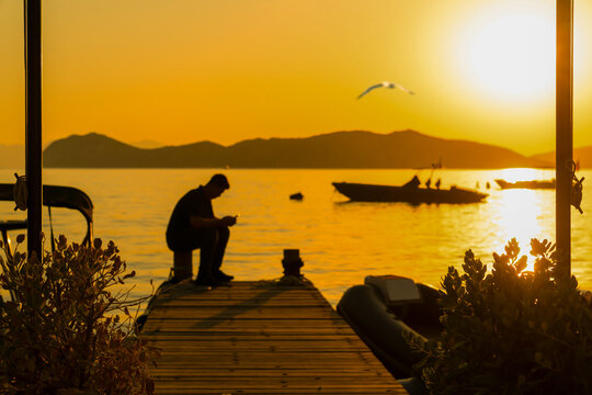 Silhouette of a lonely man using smartphone on wooden pier during golden sunset in Sogut village, Marmaris, Mugla, Turkey