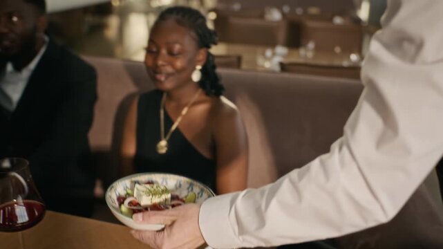 Waiter serving fresh gourmet salad to smiling African American young adult couple in upscale restaurant. Footage fits hospitality commercials and luxury dining marketing campaigns