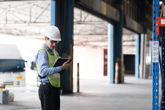 Caucasian male warehouse worker wearing white hard hat and safety vest standing in warehouse while using digital tablet manage inventory, organize logistics operations in distribution center
