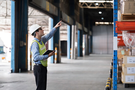 Caucasian male warehouse worker wearing white hard hat and safety vest standing in warehouse while using digital tablet manage inventory, organize logistics operations in distribution center