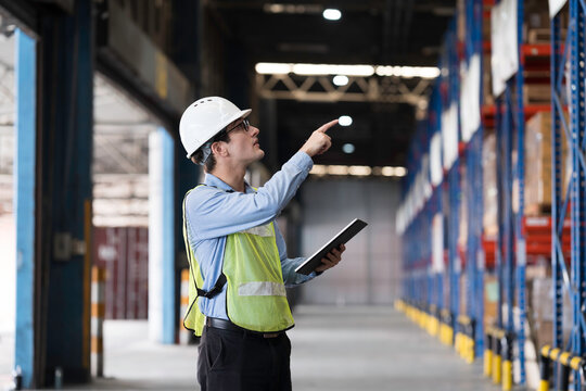 Caucasian male warehouse worker wearing white hard hat and safety vest standing in warehouse while using digital tablet manage inventory, organize logistics operations in distribution center