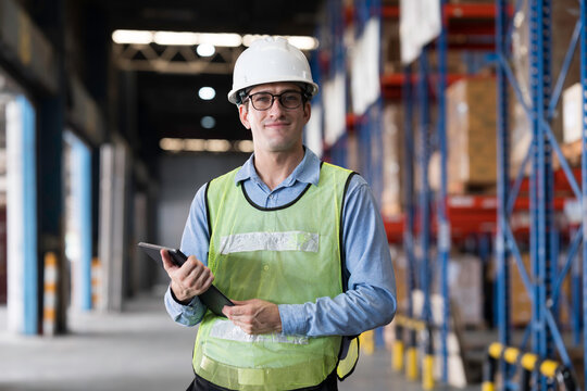 Male warehouse worker wearing white hard hat and safety vest standing in warehouse while holding digital tablet for inventory management in large distribution center