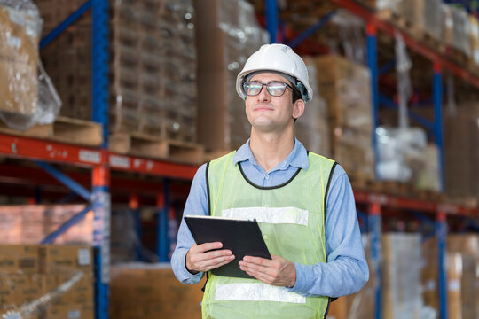 Male warehouse worker wearing white hard hat and safety vest standing in warehouse while holding digital tablet for inventory management in large distribution center
