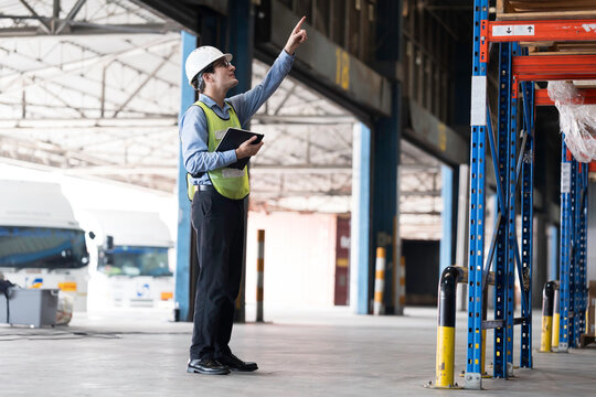 Caucasian male warehouse worker wearing white hard hat and safety vest standing in warehouse while using digital tablet manage inventory, organize logistics operations in distribution center
