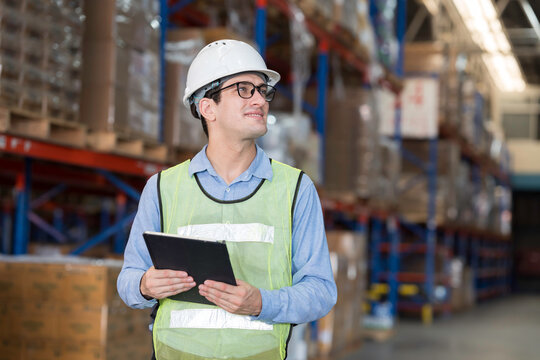 Male warehouse worker wearing white hard hat and safety vest standing in warehouse while holding digital tablet for inventory management in large distribution center
