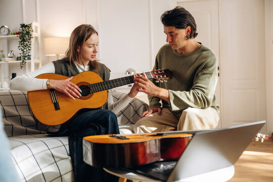 Woman playing acoustic guitar while man adjusts the strings beside her in a bright living room with a laptop and second guitar visible indoors