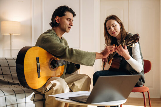 Man teaching woman to play acoustic guitar beside laptop in cozy room, informal music lesson with seated people and warm indoor lighting at home