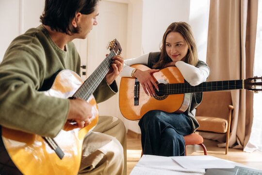 Two people with acoustic guitars sharing a relaxed indoor music session beside a window with warm natural light in a contemporary home setting