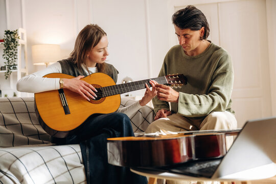 Woman playing acoustic guitar with man guiding her fingers in a cozy bright living room with reflective guitar case in foreground