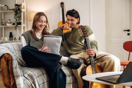 Young couple sharing a tablet while sitting on a sofa beside an acoustic guitar in a bright, comfortable contemporary living room interior