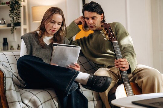 Young couple reviewing sheet music together while seated on a sofa beside an acoustic guitar in a warmly lit living room interior