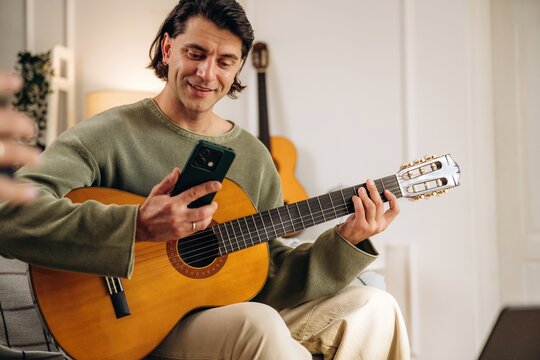 Man playing acoustic guitar while checking smartphone at home with another guitar in the background, candid indoor music practice scene