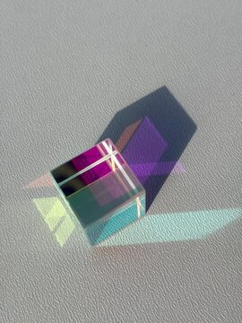 Transparent optical cube refracting colorful light into purple, turquoise and yellow patterns with geometric shadows on a textured white background.