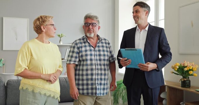 Realtor guides senior couple during apartment viewing. Footage shows agent presenting modern living room features to prospective homeowners. A homebuying decision concept for buyers.