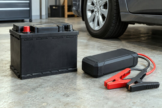 Portable car jump starter with clamps beside car battery in garage - dead battery and roadside emergency concept