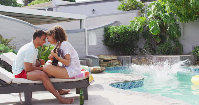 Happy diverse couple in love playing guitar, their friend jumping into swimming pool at pool party