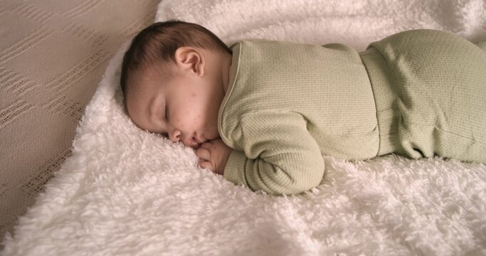 Sleeping infant lying face down on white blanket against beige cushion, wearing sage green pajamas