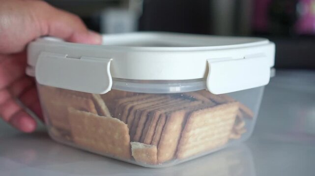 Hand placing food storage box with stacked crackers, soft natural light, Ramadan fasting time, waiting for iftar concept