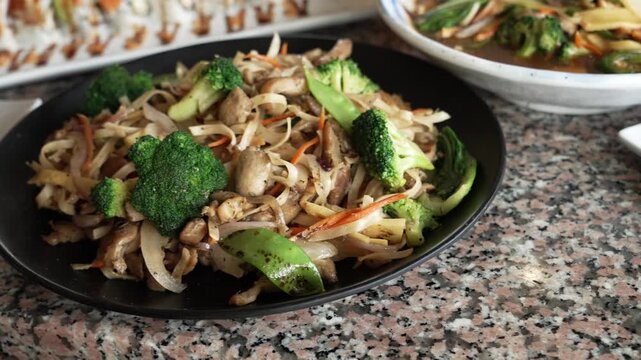 Slow motion slide reveals a vibrant plate of Pad See Ew noodles with broccoli and vegetables on granite table, surrounded by Thai cuisine dishes in a warmly lit restaurant interior.