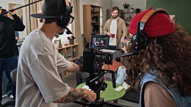 Film crew filming young adult Caucasian actors on sofa while director checking storyboard. Clip ideal for media production courses, filmmaking ads and promos