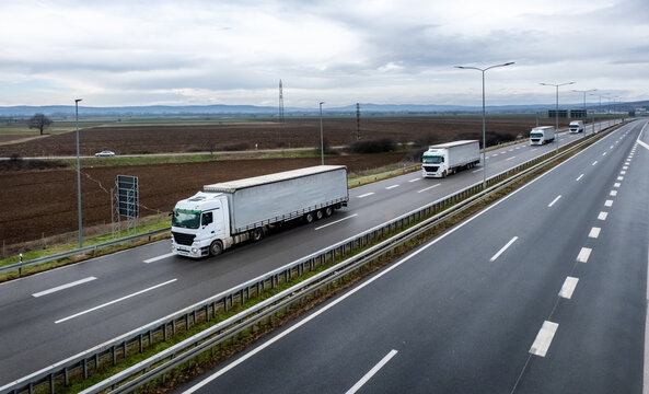 A high-angle view of multiple white semi-trailer trucks driving along a multi-lane highway through a rural landscape under an overcast sky.