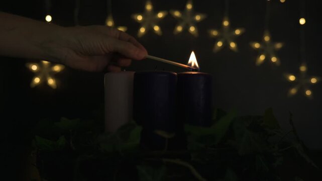 Shot of a match lighting the wick of an Advent candle. Beautiful golden bokeh background with warm string lights creating a magical Christmas preparation mood.