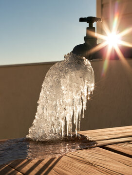 Frozen pipe burst leak beside outdoor residential spigot, thick translucent ice shell and icicle forming above wet wooden deck in bright winter sunlight