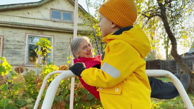 A cheerful child pushes his grandmother on a swing in a sunny garden. This slow motion shot creates a joyful, warm, and loving mood.