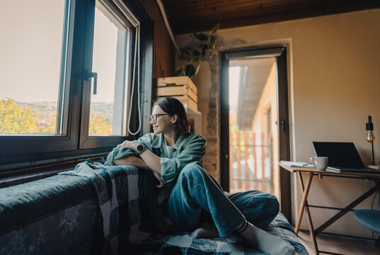 Side view profile of smiling young woman in green shirt sitting on sofa looking out window, relaxing break from remote work.