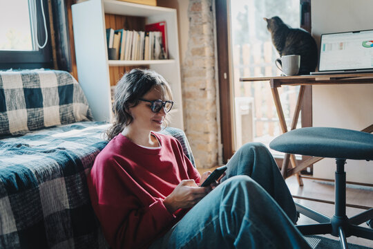 Smiling young woman in glasses looking at smartphone screen with bookshelf behind her