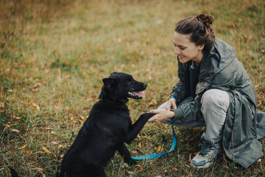 Smiling young woman in raincoat holding her black dog paw during autumn walk