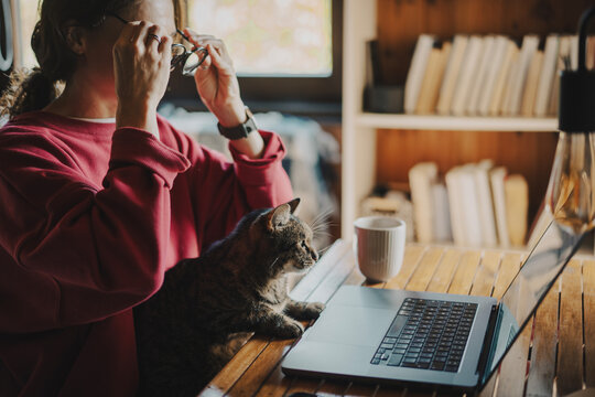 Young woman in red sweatshirt putting on glasses to start working on laptop with cat on lap