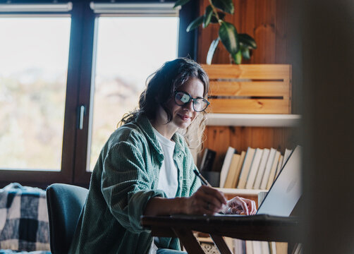 Smiling young woman around 30 years old taking notes in home office with laptop
