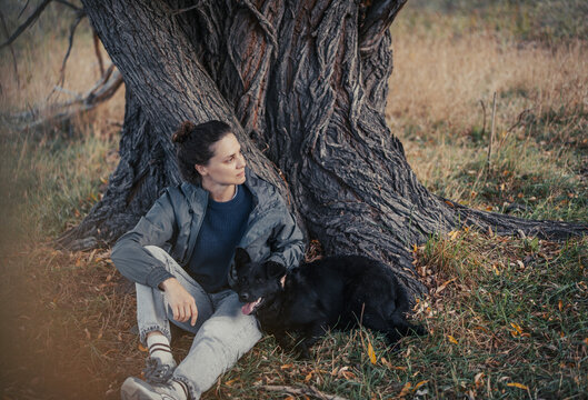 Young woman in raincoat crouching by wide willow tree trunk with her black dog on autumn overcast day