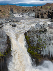 Aldeyjarfoss waterfall cascades over basalt columns in Iceland as a solitary person stands on a rocky outcrop with outstretched arms above the churning river. © Aerial Film Studio