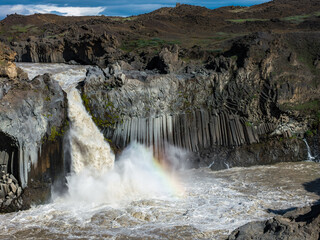 Aldeyjarfoss waterfall cascades over dark basalt columns into a turbulent pool with a faint rainbow in the northern highlands of Iceland, viewed from across the river canyon. © Aerial Film Studio