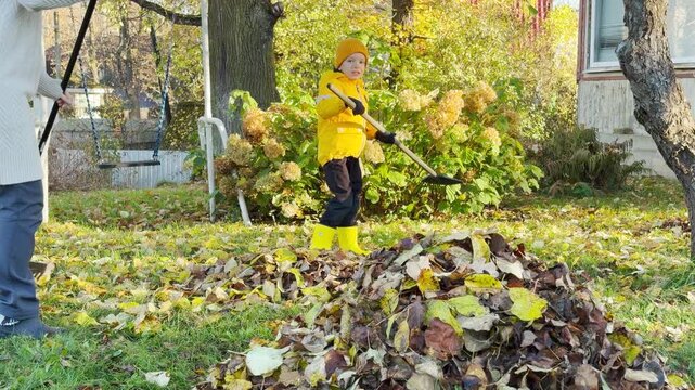 A mother and her young son rake fallen autumn leaves together into a large pile. They work joyfully in their sunlit green yard surrounded by trees. The outdoor activity fills them with warmth