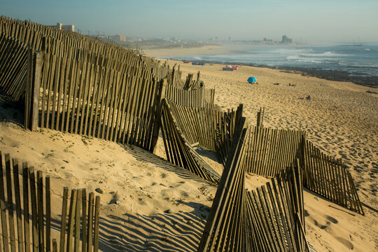 Weathered wooden dune fences casting striped shadows across a sandy beach with ocean waves and a distant city skyline