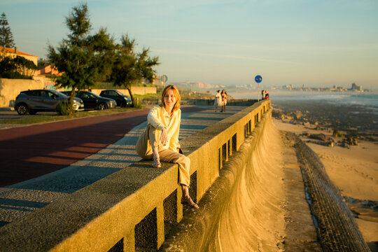 Smiling woman sitting on a seawall beside a coastal promenade and beach in warm golden evening light