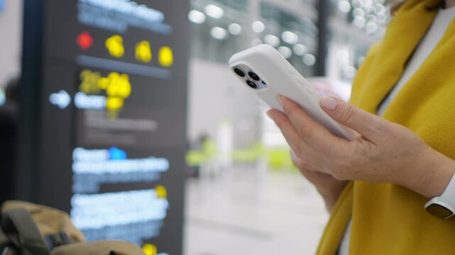 Person holds phone, looking at screen. Airport departure board visible behind. Woman wears yellow jacket. Hand holds smartphone with white case. Background shows travelers and bright lights