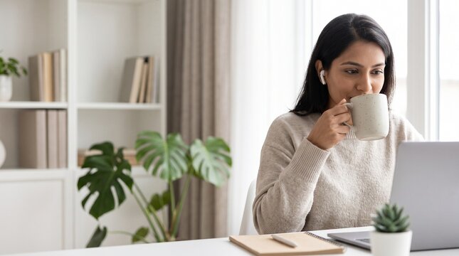 Young woman working remotely on a laptop while drinking coffee at home.