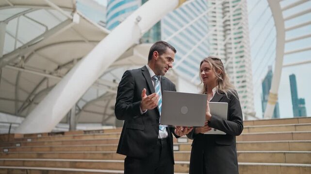 Business professionals discuss work outdoors. They stand on steps under a modern canopy. The man holds a laptop, the woman gestures. They look focused