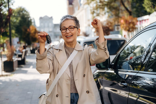Smiling older woman holding car keys with raised arms beside a black vehicle on a sunny urban street with trees, buildings, and parked cars in the background