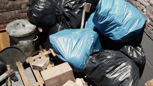 Blue and black plastic trash bags sit on a wooden pallet by a metal bin as workers prepare them for collection in the daylight.