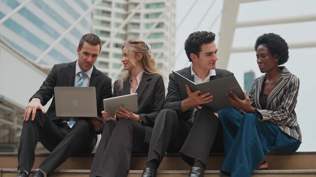Four professionals converse on urban steps. They discuss work with laptops and folders. Casual yet focused, they blend business with urban scenery