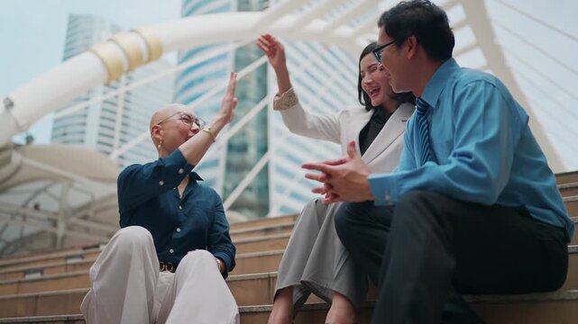 Three businesspeople celebrate together on outdoor steps. They high five with joy, then clap in approval. City buildings loom behind them