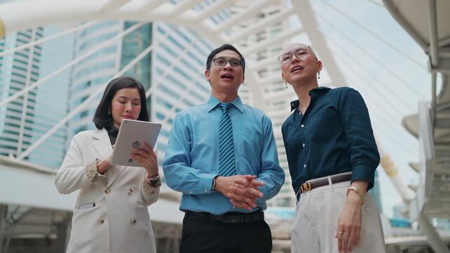 Three professionals discuss plans under a modern bridge. They gesture upward, pointing at architectural features. Woman holds a tablet, reviewing data
