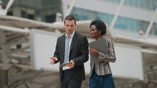 Two professionals converse while walking outdoors. They hold digital devices and papers, suggesting a meeting or review. The man gestures
