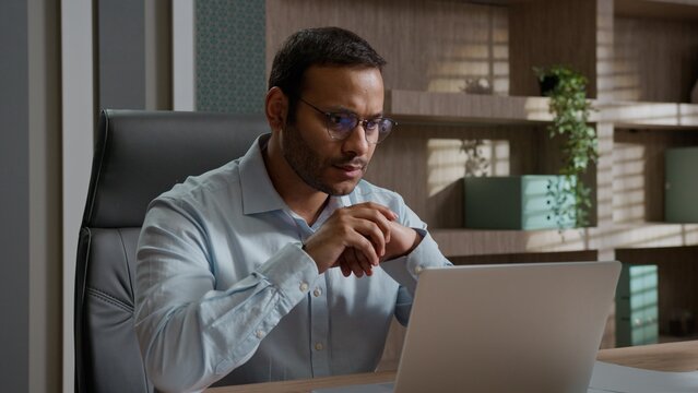 Serious focused Indian Arabian business man in glasses reading news on laptop in office worried employer programmer working workplace concentrated male businessman looking at computer screen busy job