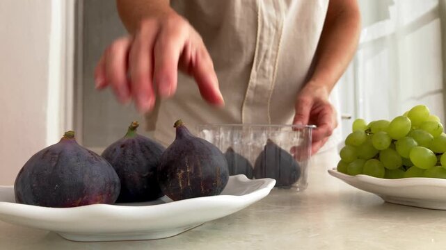 The woman carefully transfers the figs from the packaging onto a plate. Female hands place the fruit on the white plate on the table; receiving products with home delivery